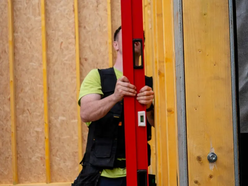 Trabajador con chaleco sujetando un nivel rojo junto a un muro de madera